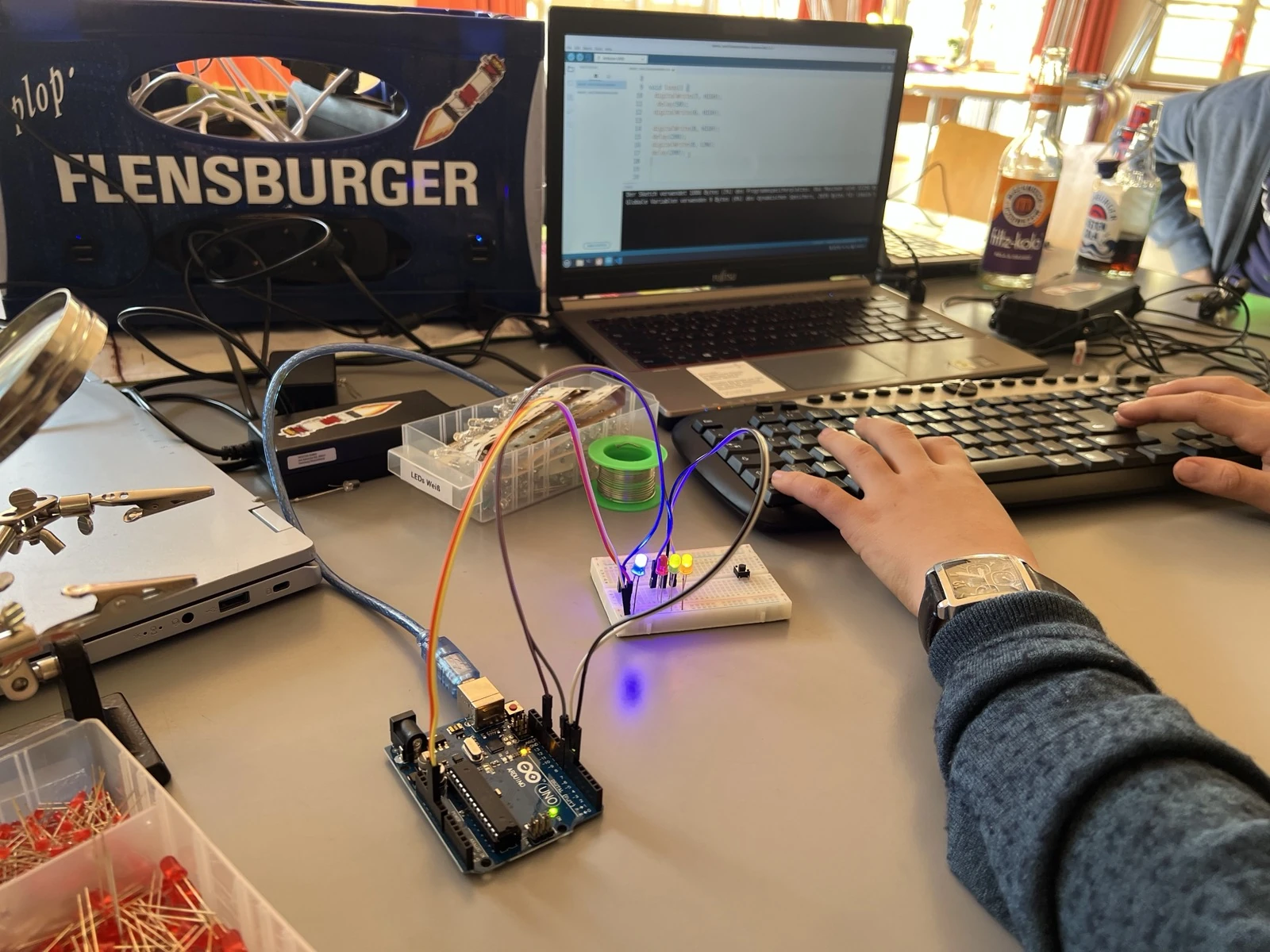 Auf einem Schreibtisch steht ein Laptop, an welchem ein Mikrocontroller mit LEDs und einem Knopf angeschlossen ist